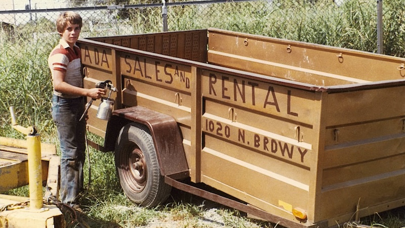 A young Wayne poses by a trailer advertising his parents' rental business.