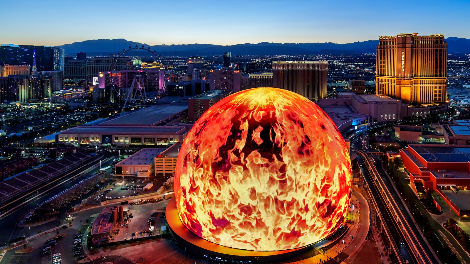 A skyline shot of Las Vegas. The Sphere in the foreground features a 360-degree outdoor screen showing fire.