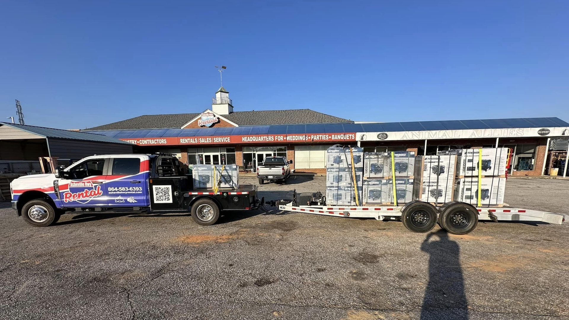 A pickup with the Asheville Highway Rental logo on its side has a trailer full of generators hitched to its trailer and is parked outside of Asheville Highway Rental.