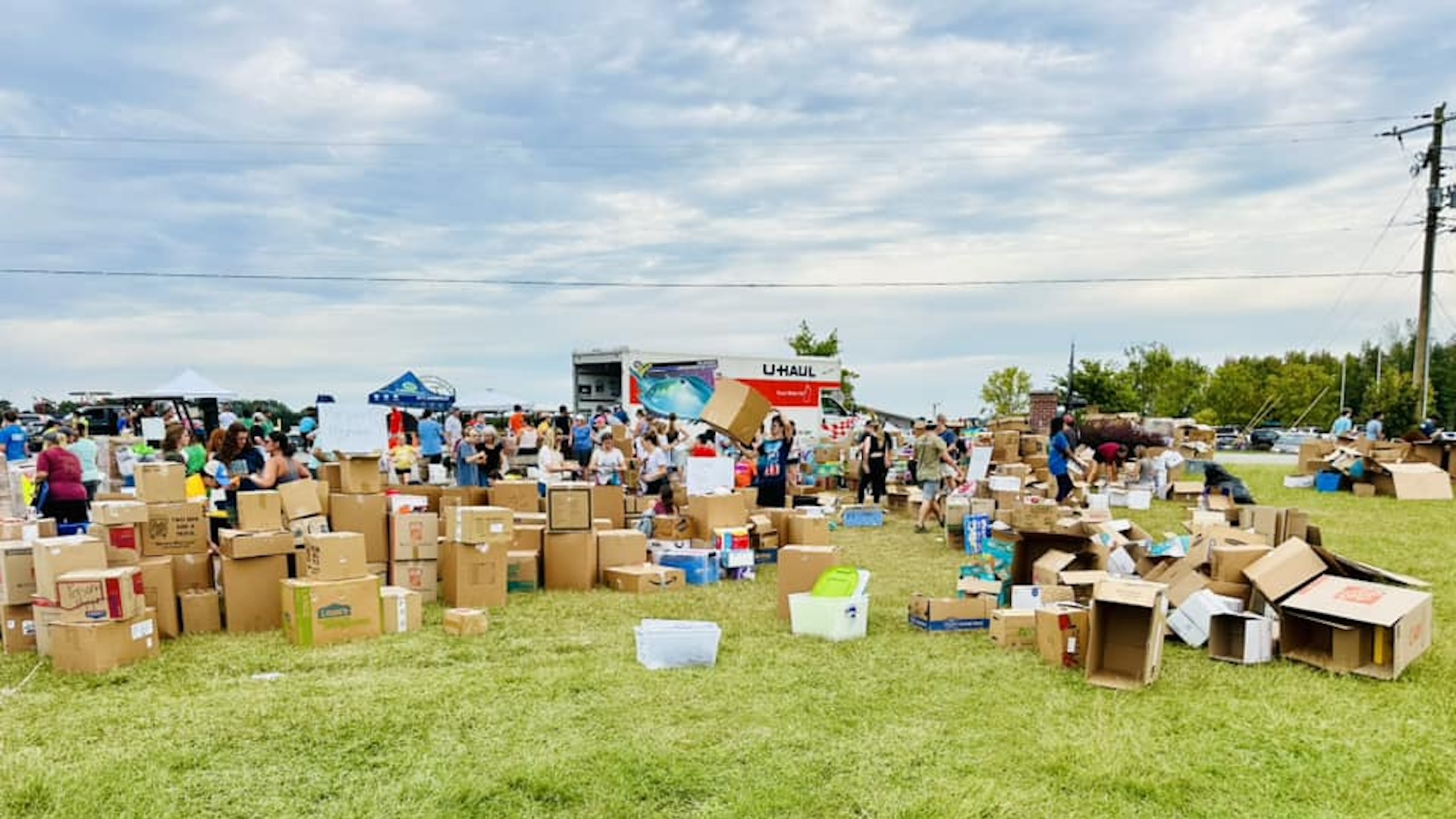 Boxes of supplies for hurricane victims are stacked and scattered in a field with a UHaul in the background.